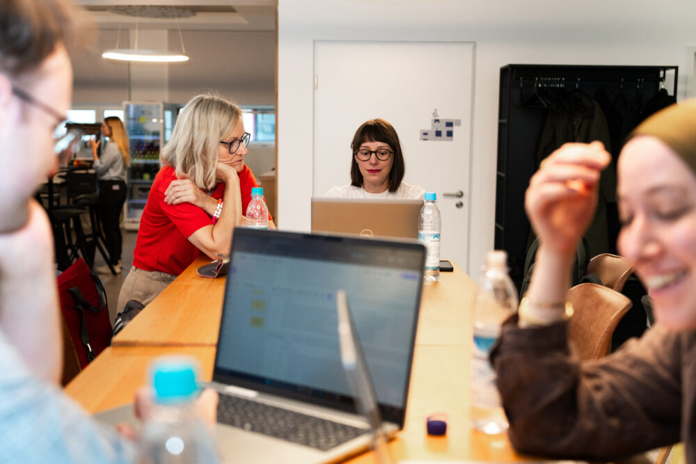 Mehrere Personen sitzen an einem Holztisch in einem modernen Raum. Sie arbeiten an Laptops, diskutieren und wirken konzentriert. Wasserflaschen und Notebooks sind auf dem Tisch verteilt. Im Hintergrund sind weitere Teilnehmer und Möbel zu sehen.