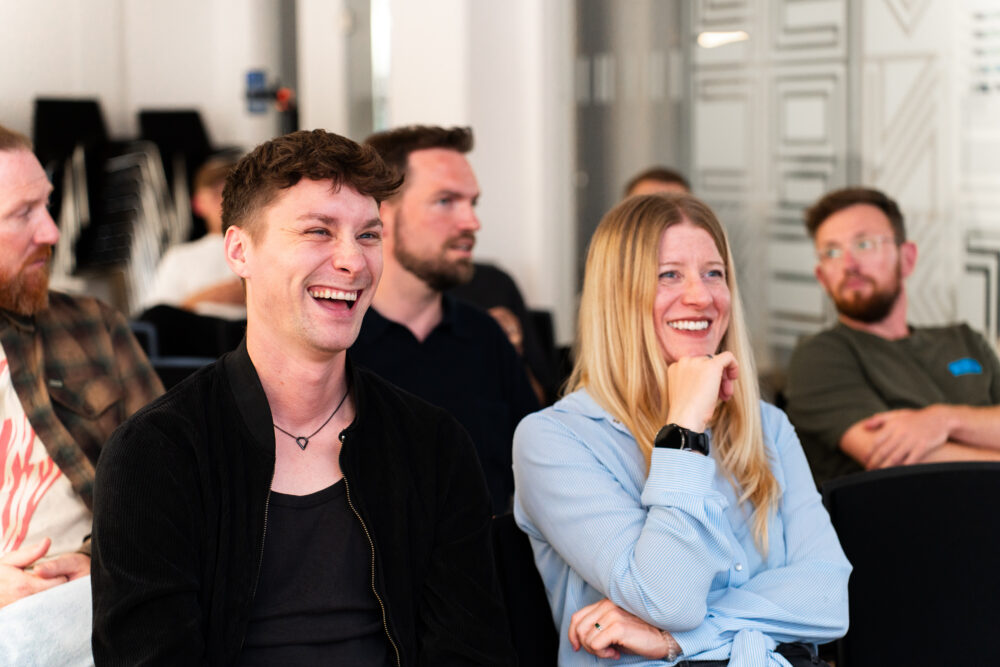 Das Bild zeigt eine Gruppe von Teilnehmern in einem Workshop. Zwei Personen im Vordergrund lachen herzlich, während andere aufmerksam zuhören. Die Umgebung wirkt modern und freundlich, mit Glaswänden und minimalistischer Einrichtung.