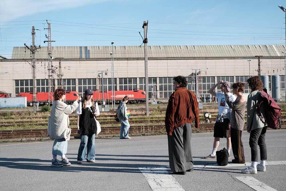 Sechs Menschen stehen neben Gleisen. Im Hintergrund ist ein Zug auf einem entfernten Gleis und eine große Industriehalle.