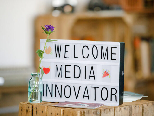 Ein Leuchtschild mit der Aufschrift Welcome Media Innovator steht neben einer Vase mit einer lilafarbenen Blume auf einem kleinen Holztisch.