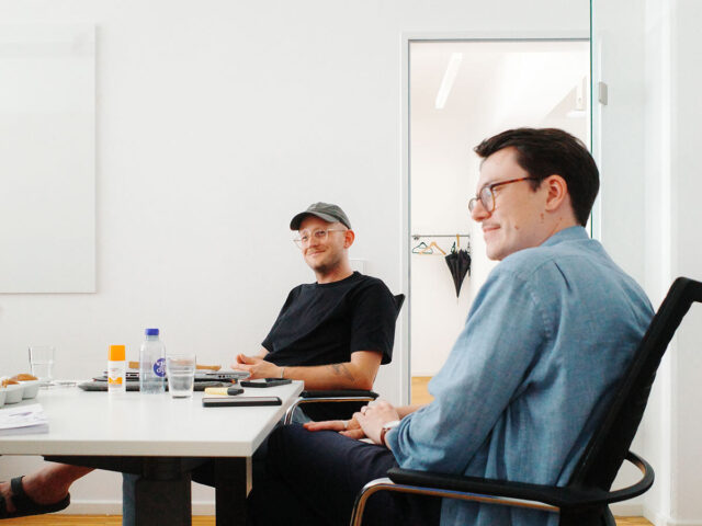 Zwei Männer sitzen in einem Büro auf Bürostühlen an einem Tisch.